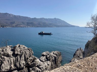 An old cargo barge with a white cabin and black hull floating on a calm blue sea, with mountains and coastal town in the background.  clipart