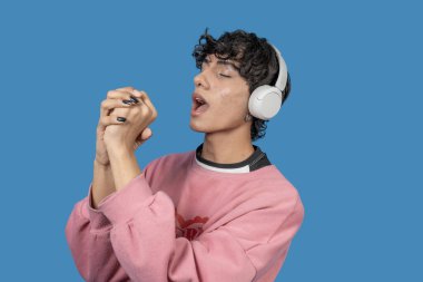 Kablosuz kulaklık takan genç latin adam mavi arka planda kapalı gözlerle şarkı söylüyor.