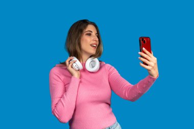 Mutlu genç bir kadın akıllı telefon ve kulaklık kullanarak selfie çekiyor, mavi stüdyo arka planında izole edilmiş.