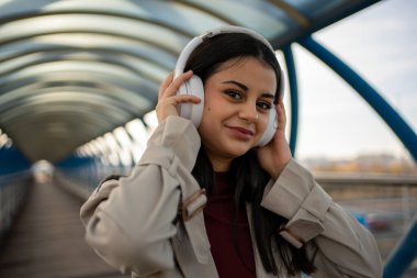 Stylish young woman enjoying music with wireless headphones while walking in a modern covered walkway clipart