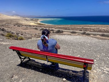 Kadın sırtüstü oturmuş, renkli bir bankta denize bakıyor.