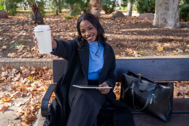 Gülümseyen iş kadını, elinde tablet tutarken ve sonbahar boyunca parkta bankta otururken kahve fincanı gösteriyor.