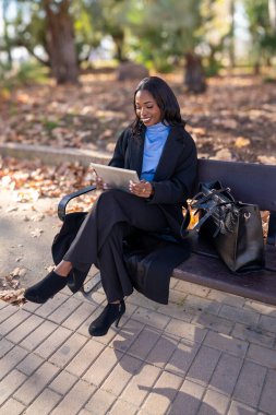 Tabletle çalışan gülümseyen iş kadını, sonbahar boyunca parkta bankta oturuyor.