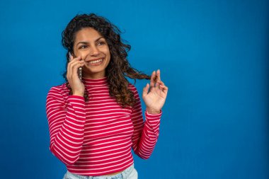 Akıllı telefondan konuşan ve mavi arka planda saçlarını kıvıran mutlu genç bir kadın.