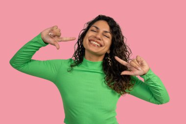 Neşeli genç bir kadın kendini işaret parmağıyla işaret ediyor, gülümsüyor ve yeşil balıkçı yaka bir gömlek giyiyor.