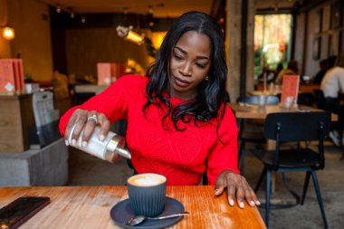 Genç siyah kadın kapuçinosuna şeker dolduruyor, modern bir kafenin ahşap masasında oturuyor.