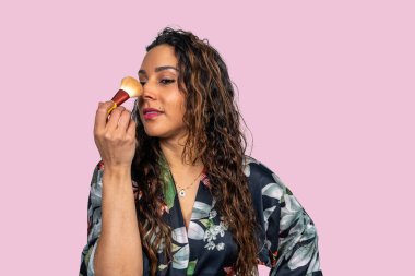 Latin bir kadın yüzüne fondöten tozu sürüyor. Pembe arka planda fırça kullanıyor.