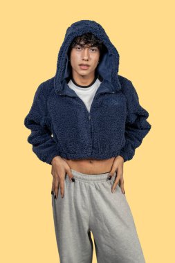 Stüdyoda mavi kürklü ceketi ve sarı arka planda gri eşofmanıyla poz veren şık Latin bir genç.