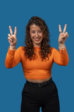 Mavi arka planda barış işareti yapan gülümseyen genç bir kadının stüdyo portresi.