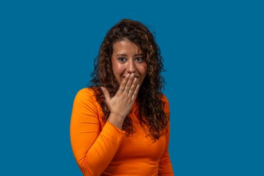 Ağzını eliyle kapatan neşeli genç bir kadının stüdyo portresi, canlı mavi bir arka planda şaşkınlığını ifade ediyor.