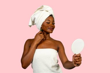 Yüzünde gua sha kazıyıcısı ile havlu takan genç bir kadın ve duştan sonra küçük bir aynaya bakıyor.