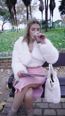 Genç bir kadın, parkta tabletini kullanırken kahveden zevk alıyor, modern yaşam tarzı ve teknolojisini sergiliyor.