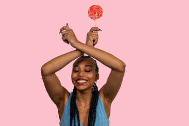 Kırmızı lolipop tutan, kollarını kavuşturup gülümseyen ve pembe arka planda duran mutlu genç kadın.