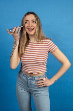 Mutlu genç kadın akıllı telefonunu diktafon olarak kullanıyor, bir sesli mesaj dinliyor ve mavi bir arkaplanda gülümsüyor.