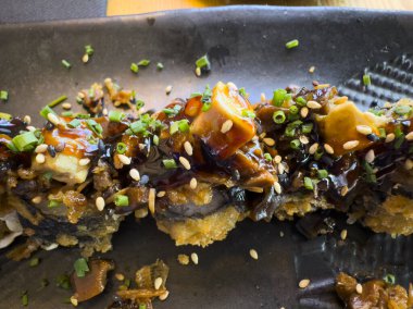 Yılan balığı, susam tohumu ve soğanla süslenmiş nefis bir tempura uramaki suşi rulosunun yakın çekimi, sade siyah bir tabakta servis edilir.