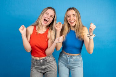 İki neşeli genç kadın sevinçlerini ve heyecanlarını, canlı mavi bir arka planda yumruklarıyla ve ağızlarını açarak ifade ediyorlar.