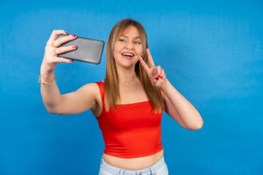Influencer mavi arka planda akıllı telefonlu selfie çekerken zafer işareti yapıyor.