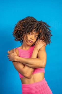Pembe bluz ve etek giyen kıvırcık saçlı genç bir kadın canlı mavi bir arkaplanda kendine sarılıyor kendini kabullenme ve vücut pozitifliğini ifade ediyor.