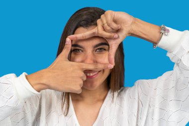 Genç bir kadın mavi arka planda parmaklarıyla yüzünü çerçeveliyor.