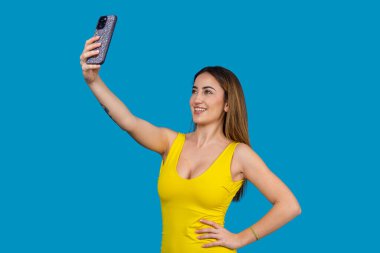 Genç bir kadın akıllı telefonuyla selfie çekiyor, gülümsüyor ve mavi bir arka planda poz veriyor.
