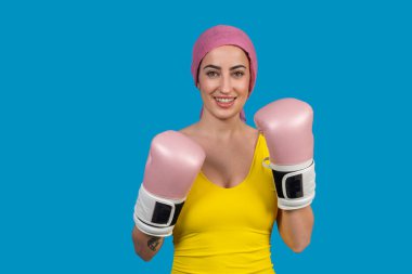 Pembe boks eldiveni ve başörtüsü takan gülümseyen genç bir kadının stüdyo portresi. Kanserle pozitif tutumla savaşıyor.