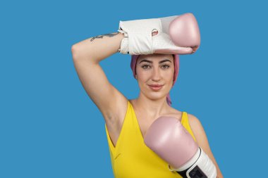 Pembe boks eldiveni ve başörtüsü takan genç bir kadının stüdyo portresi, canlı mavi bir arka plana karşı poz veriyor.