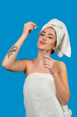 Havluya sarılı genç bir kadın duş aldıktan sonra yüzüne güzellik serumu sürüyor.