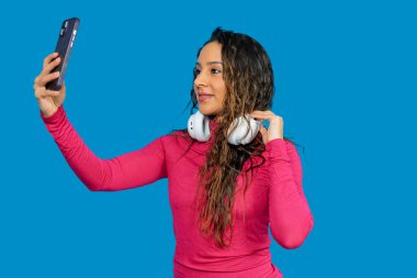 Genç Latin bir kadın mavi arka planda akıllı telefon ve kulaklıkla selfie çekiyor.