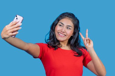 Mutlu genç bir kadın, mavi stüdyo arka planında izole bir şekilde zafer işareti yaparken selfie çekiyor.