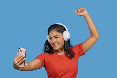Kulaklıkla müzik dinleyen mutlu Latin kadın selfie çekiyor ve mavi arka planda dans ediyor.