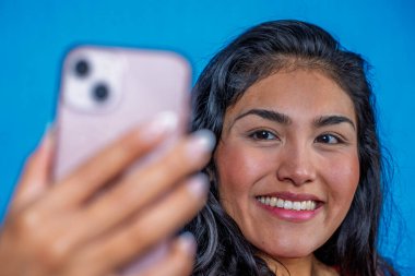 Neşeli İspanyol kadın mavi arka planda cep telefonuyla selfie çekiyor.