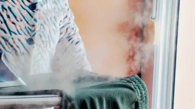 Ev hanımı ütü masasındaki yeşil pantolonları ütülüyor, buharlı ütü kullanıyor.