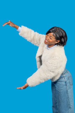 Modaya uygun genç bir kadın elleriyle görünmez bir şey tutuyor gibi, mavi bir arka planda gülümsüyor.