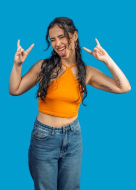 Kaygısız genç kadın mavi arka planda dikilirken dilini çıkarıp rock 'n roll hareketi yapıyor.