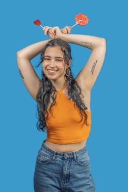 Canlı, mavi bir arka planda iki lolipop, bir kalp ve bir girdap tutan neşeli genç bir kadının stüdyo portresi.