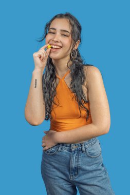 Uzun siyah saçlı, sarı şekerleme yiyen ve mavi arka planda gülümseyen mutlu bir genç kadın.
