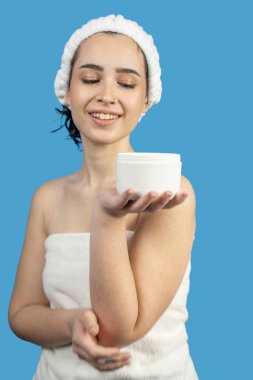 Duştan sonra elinde bir kavanoz nemlendirici krem tutan genç bir kadının güzellik rutini, mavi arka planda izole edilmiş.
