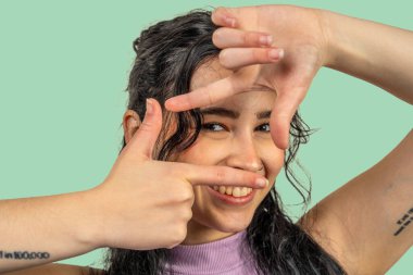 Neşeli genç bir kadın, parmaklarıyla yüzünü çerçeveliyor, gülümsüyor ve canlı yeşil bir arka plana karşı anın tadını çıkarıyor.