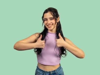 Mutlu genç bir kadın iki eliyle baş parmak işareti yapıyor. Olumlu ve yeşil bir arkaplan üzerinde mutabık.