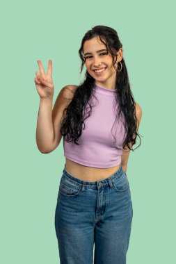 Neşeli genç bir kadın zafer işaretini iki parmakla gösteriyor, gülümsüyor ve canlı yeşil bir arka planda pozitifliğini gösteriyor.