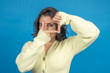 Genç bir kadın, parmaklarıyla parlak mavi bir arkaplanda bir çerçeve yapıyor.