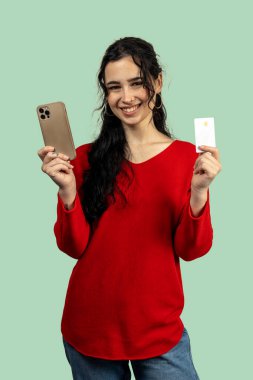 Mutlu genç bir kadın elinde akıllı telefon ve kredi kartı tutuyor. İnternetten alışveriş ve güvenli ödeme öneriyor.