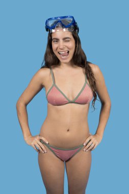Bikini giyen, dalış maskesi takan, mavi arka planda dilini sallayan genç bir kadın.