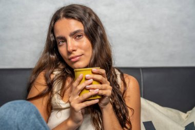 Huzurlu genç bayan, kanepede sessiz bir anın tadını çıkarıyor, elinde sıcak bir kupa tutuyor.