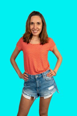 Canlı bir turkuaz arka planda elleri kalçasında poz veren neşeli genç bir kadının stüdyo portresi.