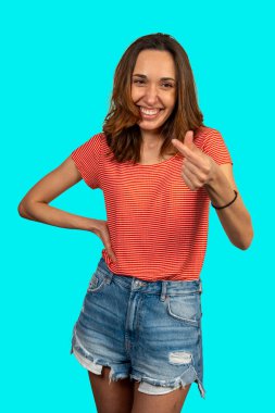 Küçük bir kalp işareti yapan, gülümseyen ve turkuaz bir arka planda poz veren neşeli genç bir kadının stüdyo çekimi.