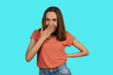 Turkuaz bir arka planda gülerken ağzını eliyle kapatan neşeli genç bir kadının stüdyo portresi.