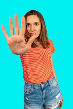 Elinde turkuaz bir arka planda, kendine güvenini ve kendine güvenini ifade eden dur hareketi yapan genç bir kadın.