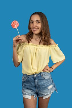 Kırmızı beyaz bir lolipop tutan neşeli genç bir kadının canlı mavi bir arka planda izole edilmiş bir fotoğrafı.