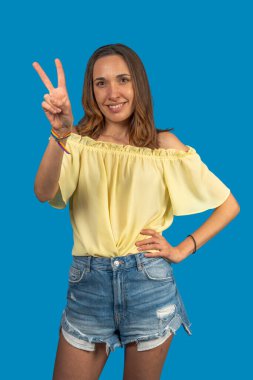 Sarı bluz ve kot pantolon giyen beyaz kadın mavi arka planda gökkuşağı bileziği olan zafer işareti gösteriyor.